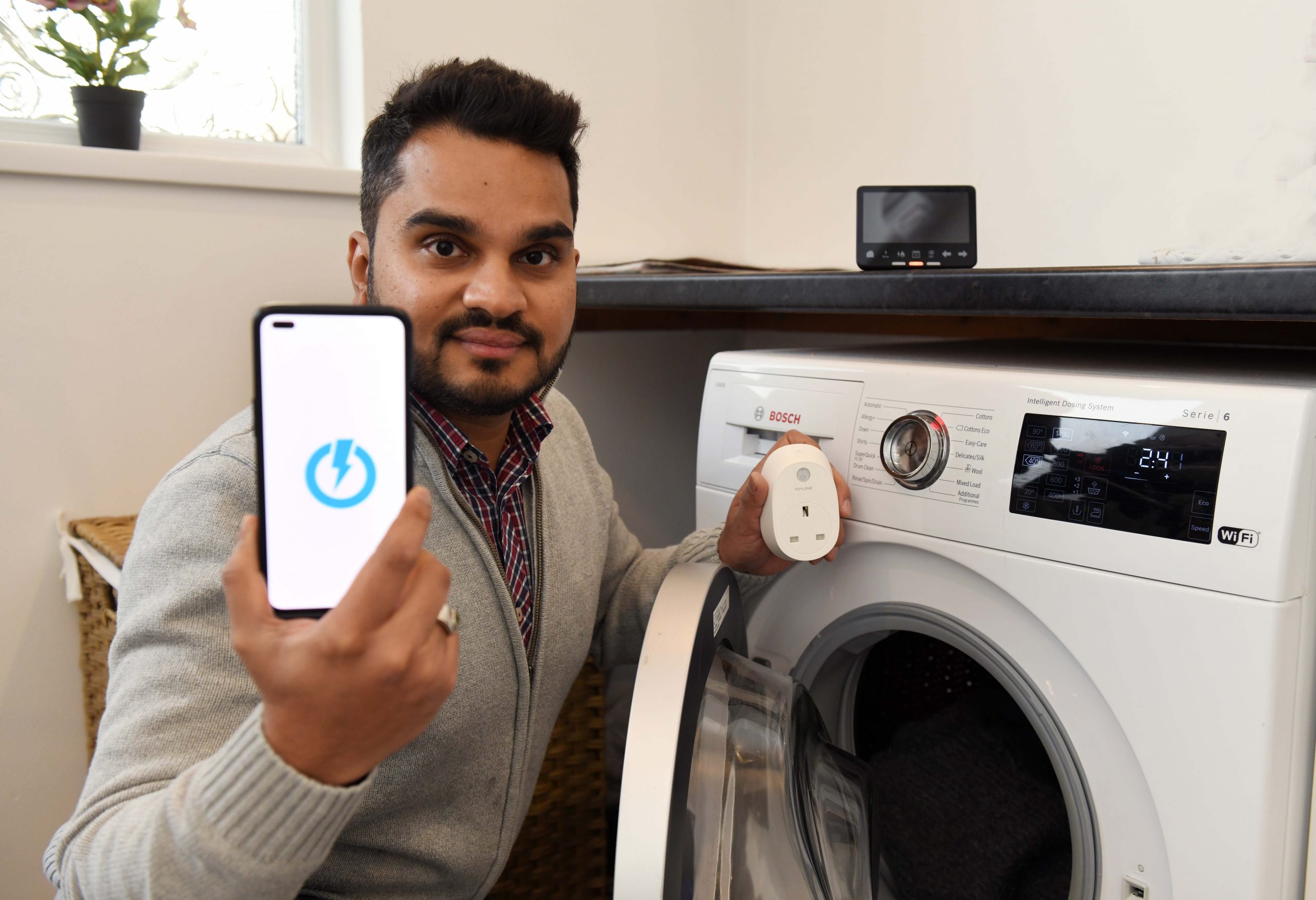 A man crouches in front of a washing machine, holding a smartphone showing the equiwatt app and a smart plug.
